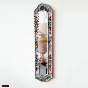 Peut inclure: Un miroir étroit et haut avec un cadre décoratif. Le cadre est en bois peint en blanc et présente un motif floral avec des fleurs roses, bleues et vertes. Le miroir a un bord biseauté et un sommet légèrement arqué.