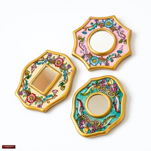 May include: Three decorative mirrors with floral designs. Each mirror has a different shape and color scheme: cream with red flowers, pink with blue flowers, and teal with yellow flowers. All have gold-colored frames.