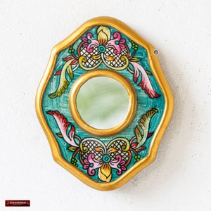 May include: A decorative wall mirror with a gold-painted frame and a teal background. The mirror has a central circular mirror and is adorned with colorful floral and scroll designs in pink, yellow, and green.