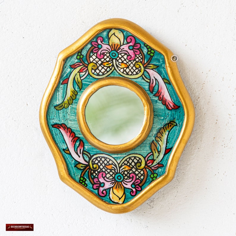 May include: A decorative wall mirror with a gold-painted frame and a teal background. The mirror has a central circular mirror and is adorned with colorful floral and scroll designs in pink, yellow, and green.