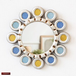 May include: A round decorative mirror with a silver-colored frame and a sunburst design. The frame features alternating blue and yellow glitter-filled circles. The mirror is mounted on a white wall, reflecting a room's interior.