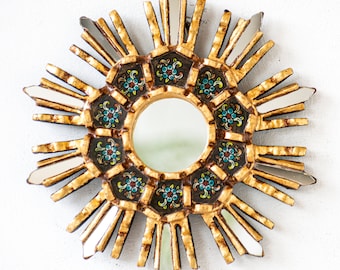 May include: A gold sunburst mirror with a round, mirrored centre and a black and gold frame decorated with colourful floral designs.