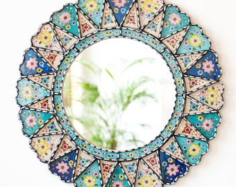 May include: A round mirror with a decorative frame made of blue, green, yellow, and pink floral tiles. The mirror has a scalloped edge.