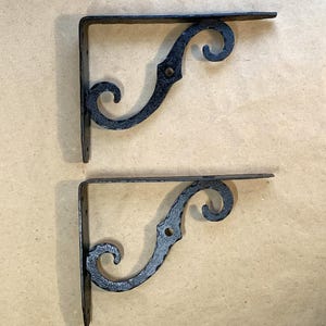 May include: Two black, wrought iron shelf brackets. Each bracket has a right angle shape with a flat top and a decorative, curved scroll design. The brackets are made of a textured metal and are designed for wall mounting.