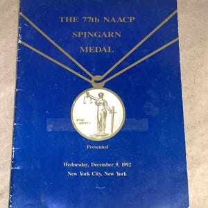 May include: A blue pamphlet with gold lettering announcing the 77th NAACP Spingarn Medal presented on Wednesday, December 9, 1992 in New York City, New York. The cover features a gold illustration of a medal with a woman holding scales of justice and a sword.