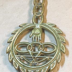 May include: A brass trivet with a hand holding a heart in the center. The trivet is surrounded by a wreath of leaves and has a chain hanging from the top.