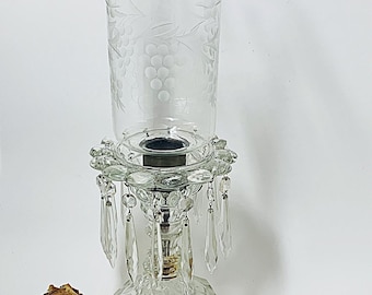 May include: A clear glass candle holder with a decorative grape design on the top. The base is a faceted design with hanging crystal accents. A dried rose is in the foreground.