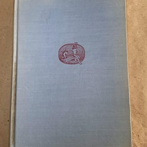 May include: A light blue hardcover book with a red oval emblem. The emblem features a figure with a bow and arrow. The book's cover has a textured appearance.