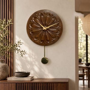 May include: A circular wall clock featuring a dark brown wood grain finish with a carved floral petal pattern. The clock face includes thin gold-coloured hands and a matching gold-coloured pendulum with a circular weight hanging from the centre.