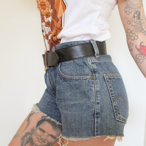 May include: A person wearing a white t-shirt with a brown and orange graphic print, a black leather belt, and denim shorts with frayed edges.