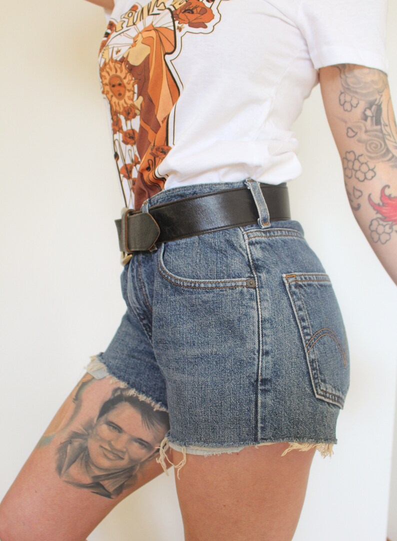 May include: A person wearing a white t-shirt with a brown and orange graphic print, a black leather belt, and denim shorts with frayed edges.