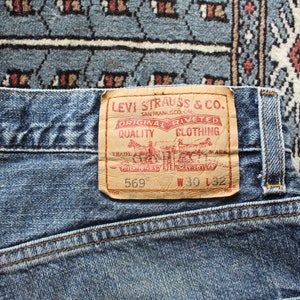 May include: A close-up of a blue denim jean with a brown leather Levi Strauss & Co. patch. The patch has red text that reads "Original  Riveted Quality Clothing" and the text "569" and "W 30 L 32".
