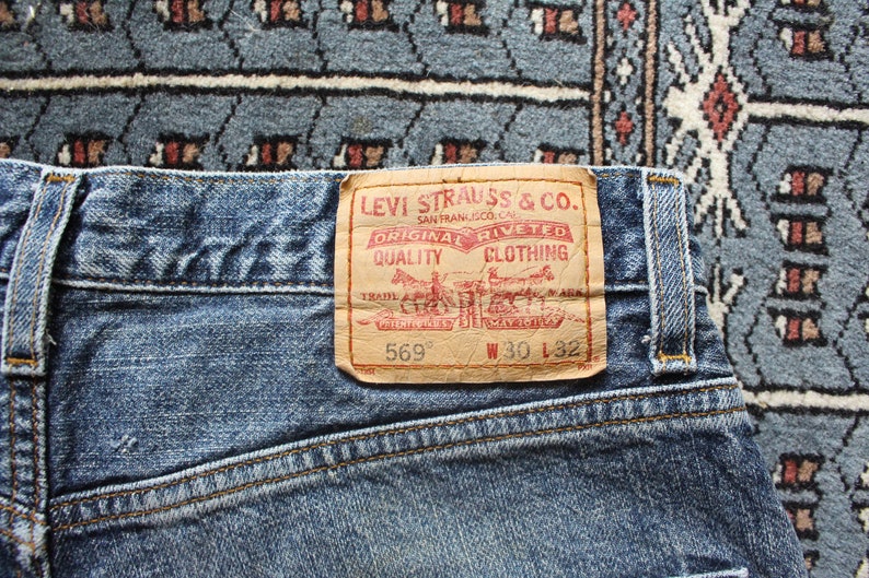 May include: A close-up of a blue denim jean with a brown leather Levi Strauss & Co. patch. The patch has red text that reads "Original  Riveted Quality Clothing" and the text "569" and "W 30 L 32".