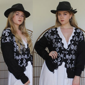 May include: A black crochet cardigan with a long sleeve and a V-neck. The cardigan is worn over a white dress and features a delicate floral pattern.