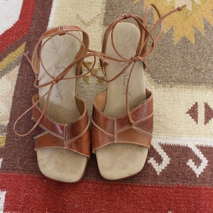 Puede incluir: Un par de sandalias de cuero marrón con cierres de encaje. Las sandalias tienen un diseño tejido y el nombre de la marca "Sun Rivers" es visible en la plantilla.