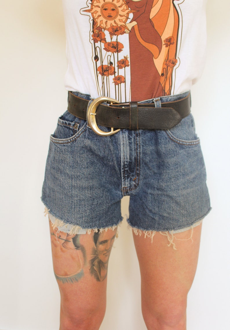 May include: A pair of blue denim shorts with frayed edges. The shorts are worn with a brown leather belt with a gold buckle.
