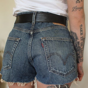 May include: A person wearing a white t-shirt with a small anchor tattoo and the words "S-H Laugh" on their arm. They are also wearing a black belt and denim shorts with frayed edges. The person has a tattoo on their leg that says "Take it or leave it."