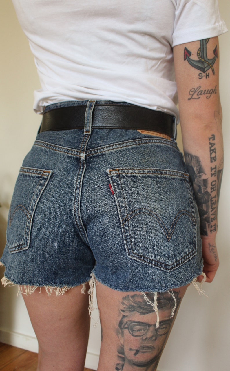 May include: A person wearing a white t-shirt with a small anchor tattoo and the words "S-H Laugh" on their arm. They are also wearing a black belt and denim shorts with frayed edges. The person has a tattoo on their leg that says "Take it or leave it."