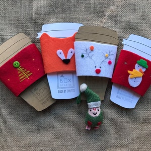 May include: Four felt coffee cup sleeves with different holiday designs. The sleeves are red, orange, white, and green. The designs include a Christmas tree, a fox, a reindeer, and a snowman. The sleeves are made by The Box Stories and say "Made by Shruette".