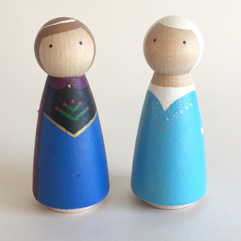 May include: Two wooden peg dolls dressed as the characters from the movie Frozen. The doll on the left is dressed in a blue and purple dress with a black bodice and a green and red design. The doll on the right is dressed in a light blue dress with white accents.