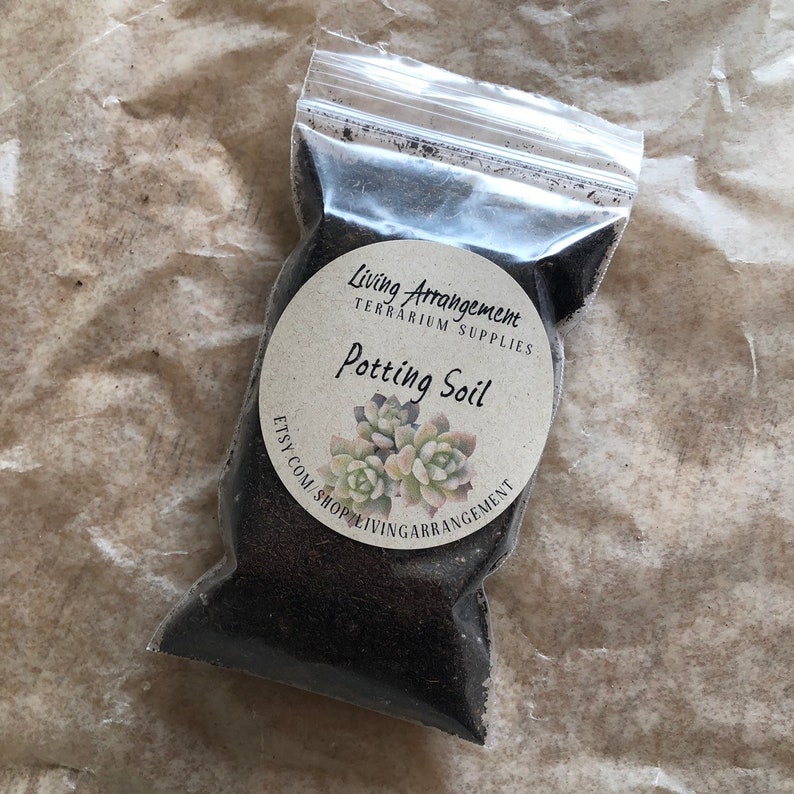 May include: A clear plastic bag filled with dark brown potting soil. The bag has a white circular label that reads "Living Arrangement Terrarium Supplies Potting Soil" with a small succulent plant illustration.