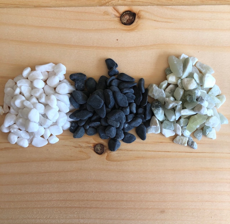 May include: Three piles of decorative stones, one white, one black, and one green. The stones are arranged in a row on a wooden surface.
