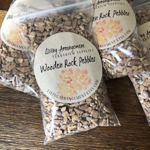 May include: A bag of small, brown, wooden rock pebbles for terrariums. The bag has a label that says "Living Arrangement Terrarium Supplies Wooden Rock Pebbles" and includes the website address "livingarrangement.etsy.com".