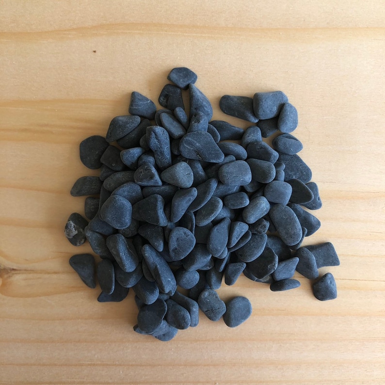 May include: A pile of smooth, dark gray river rocks. The rocks are various sizes and shapes, and they are arranged in a loose pile.