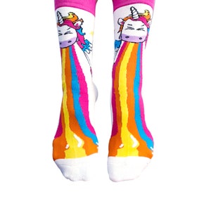 May include: A pair of white socks with a pink cuff. The socks feature a cartoon unicorn with a rainbow coming out of its mouth. The rainbow is made up of yellow, orange, red, blue, and green stripes.