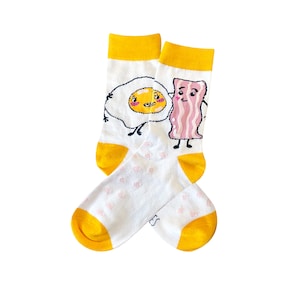 May include: A pair of white socks with yellow cuffs. One sock features a cartoon egg with a smiling face and the other sock features a cartoon strip of bacon with a smiling face.