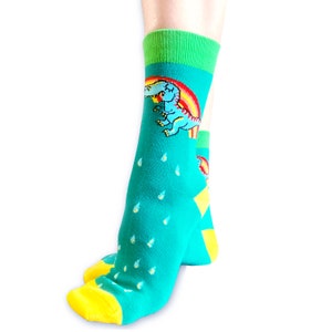 May include: A pair of teal blue ankle socks with a yellow and green trim. The socks feature a cartoon dinosaur eating a rainbow.
