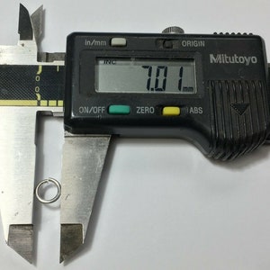 May include: A silver metal ring being measured with a digital caliper. The caliper reads 7.01 mm.