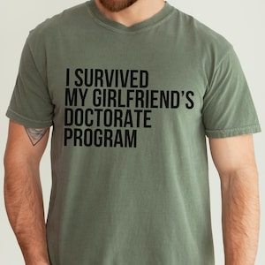 May include: A green t-shirt with the text "I survived my girlfriend's doctorate program" printed in black.