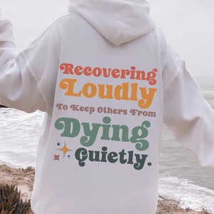 May include: White hooded sweatshirt with the text "Recovering Loudly To Keep Others From Dying Quietly." in rainbow colors.