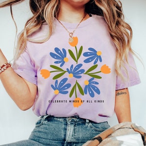 May include: A lavender t-shirt featuring a floral design in shades of blue and orange. The text "Celebrate Minds of All Kinds" is printed on the shirt.