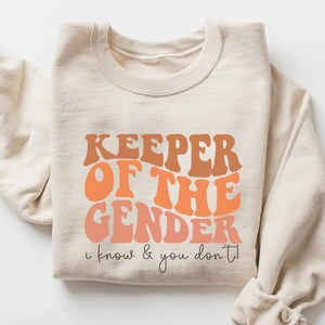 May include: A beige sweatshirt with the text "KEEPER OF THE GENDER I know & you don't!" printed in orange and brown.