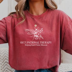 May include: A maroon t-shirt with the words "OCCUPATIONAL THERAPY Helping Each Flower Blossom" and a white graphic of a bee with floral accents. The shirt is a relaxed fit.