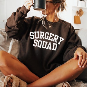 May include: A woman wearing a brown sweatshirt with the text "SURGERY SQUAD" in white letters. She is sitting on a sofa and holding a black mug.