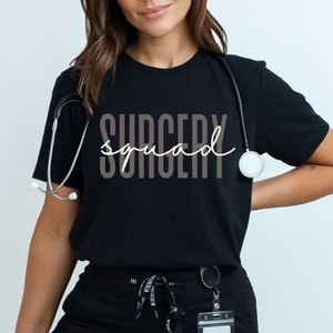Op de afbeelding: Een zwart T-shirt met de tekst "Surgery Squad" in lichtbruine kleur. Het shirt wordt gedragen door een persoon met een stethoscoop om de nek.