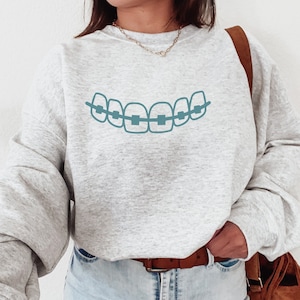 May include: A gray sweatshirt with a blue graphic of a mouth with braces. The sweatshirt has a relaxed fit and is worn by a person with long brown hair. The person is wearing blue jeans and a brown belt.