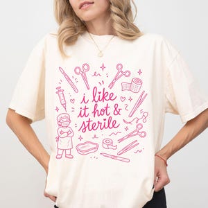May include: An ivory t-shirt featuring a pink graphic print of medical instruments, a surgical figure, and the text "i like it hot & sterile". The design includes scissors, a syringe, a scalpel, and bandages arranged around the central text.