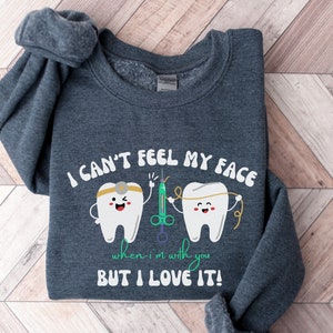 May include: A dark gray sweatshirt with the text "I CAN'T FEEL MY FACE" and the phrase "when I'm with you BUT I LOVE IT!". The graphic features two cartoon teeth, a syringe, and dental floss. The sweatshirt is folded.