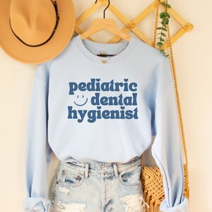 May include: Light blue sweatshirt with the text "pediatric dental hygienist" in blue with a smiley face and a heart. The sweatshirt is on a hanger with a straw hat and a woven bag.