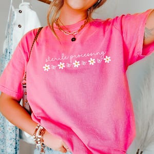 May include: A bright pink short-sleeve t-shirt featuring the white text sterile processing centered above a row of white daisies and small butterfly outlines. The shirt has a crew neckline and a relaxed fit, showcasing a vibrant solid color.