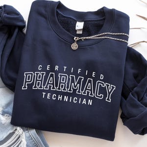 May include: A navy blue crewneck sweatshirt featuring the white text "CERTIFIED PHARMACY TECHNICIAN" in a collegiate-style font. The garment has long sleeves and a ribbed neckline, with a silver-toned chain necklace resting on the collar area.