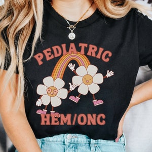 Peut inclure: T-shirt noir avec un arc-en-ciel et deux fleurs de dessin anim&eacute;. Le texte "PEDIATRIC HEM/ONC" est imprim&eacute; sur le t-shirt.