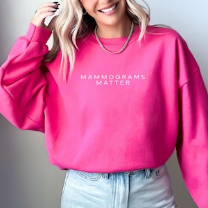 May include: A bright hot pink crewneck sweatshirt featuring the white text MAMMOGRAMS MATTER printed across the chest. The garment has long sleeves with ribbed cuffs and a ribbed neckline, providing a simple and clean design for this casual top.