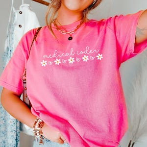 May include: A bright pink short-sleeve t-shirt featuring the white text medical coder written in a cursive script, accompanied by a row of white daisy flowers and small butterfly silhouettes printed across the center of the chest area.