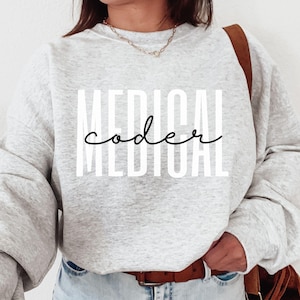 May include: An ash grey crewneck sweatshirt featuring the white block-lettered text MEDICAL and the black cursive text coder layered over it. The garment has long sleeves and a ribbed neckline, providing a neutral base for the graphic design.