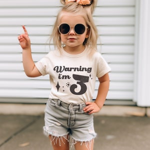 May include: A young child wearing a light beige t-shirt with the text "Warning I'm 3" in black. The child is wearing round sunglasses and has their hair pulled back in two pigtails with a pink flower.